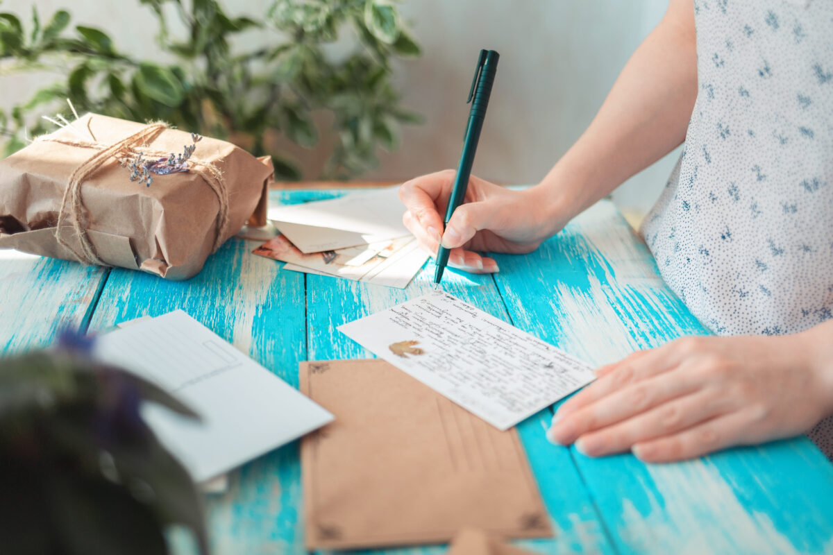 A woman writes a letter, next to it is a craft envelope. Hands lose-up. Blue wooden table with a parcel in the background. The concept of postcrossing.