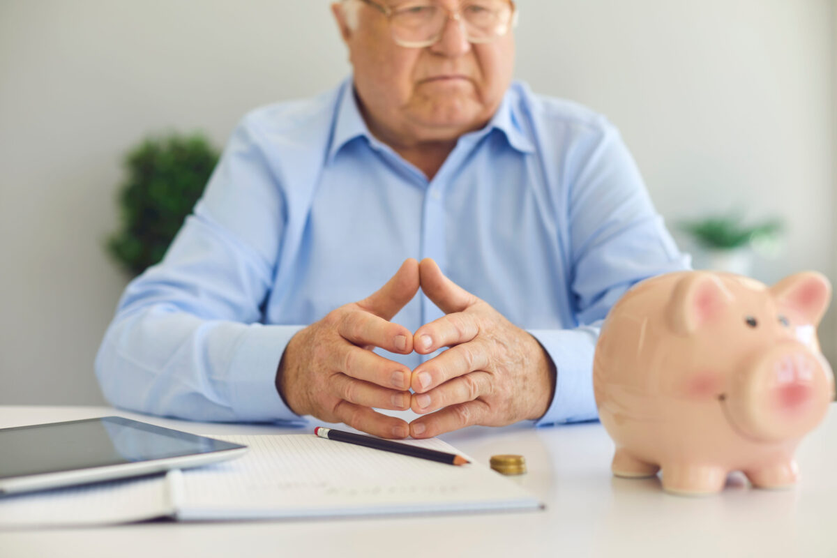 Serious senior man sitting at desk with notebook, tablet computer and piggy bank, planning budget, managing personal finance, thinking over pension plan or monthly annuity payment. Hands in soft focus