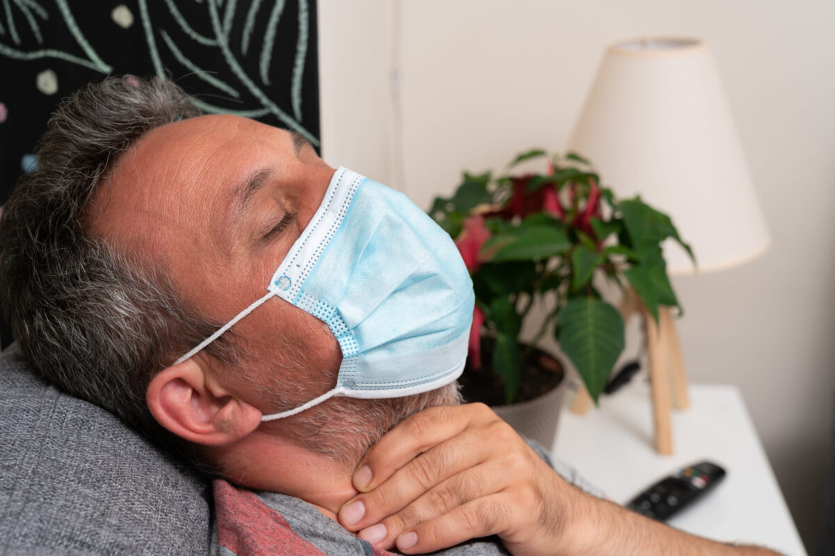Close-up of ill adult man having cold flu influenza sars covid19 symptoms touching neck with sore throat wearing protective medical or surgical disposable mask at home