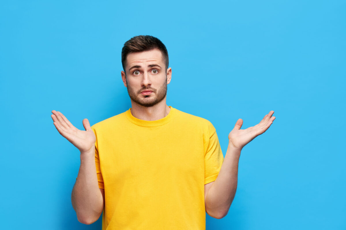Young confused man spreads hands sideways feels doubt while makes choice