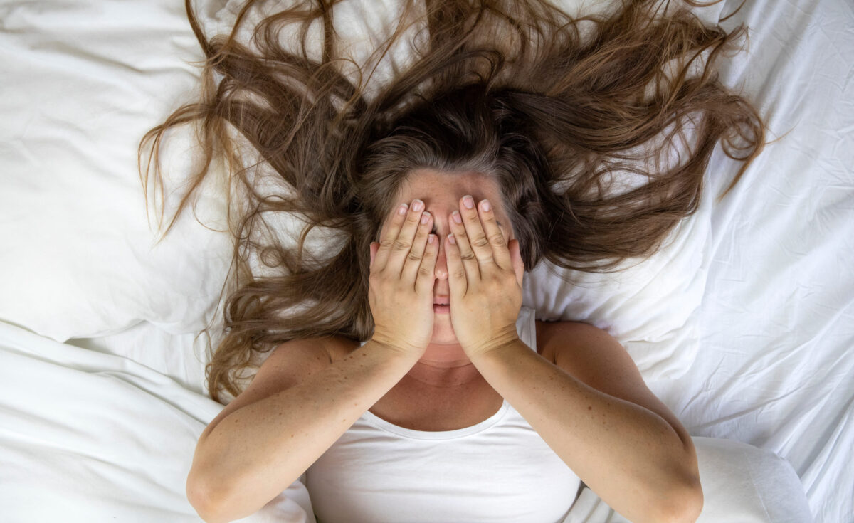 Young woman lying in bed suffering, tired woman covering face with hands, can't sleep feels exhaustand on white sheets in bedroom closeup