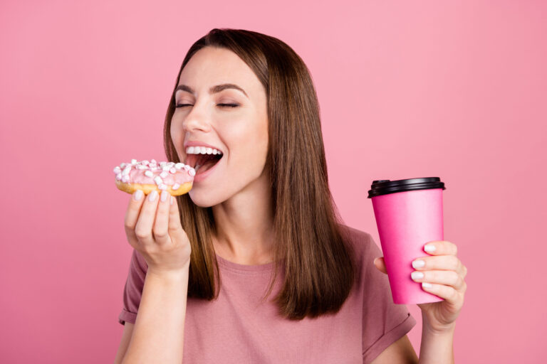 Portrait of attractive dreamy cheerful girl eating doughnut drinking latte isolated over pink pastel color background