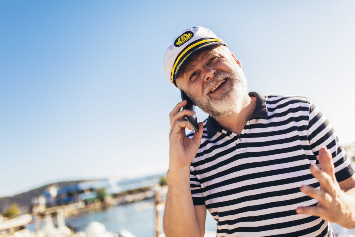 Mature man standing near the sea dressed in a sailor's shirt and hat using smart phone