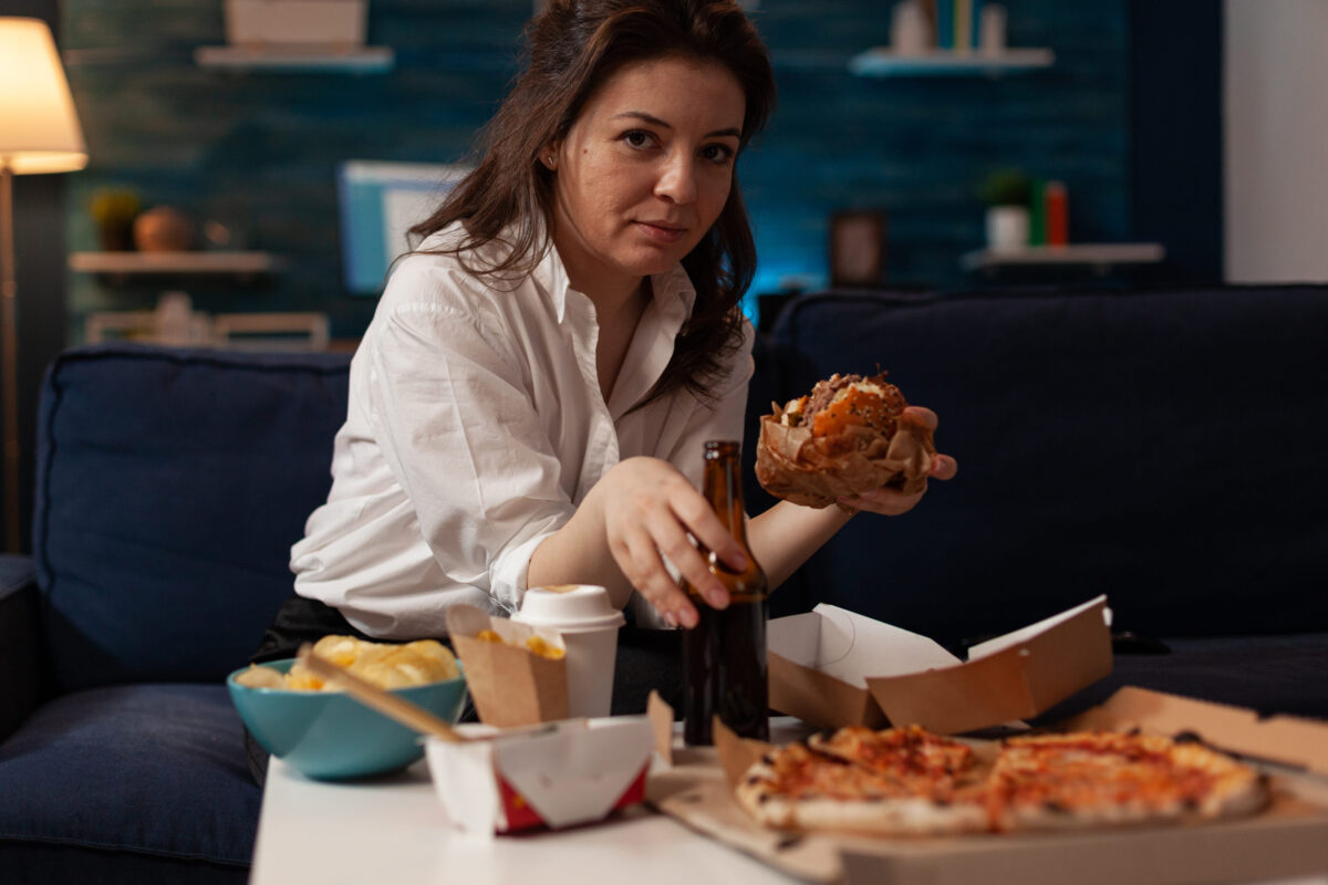 Woman holding beer bottle eating tasty takeaway hamburger while relaxing on couch in front of tv table with fast food menu. Hungry person enjoying junk food sitting on sofa in the evening.