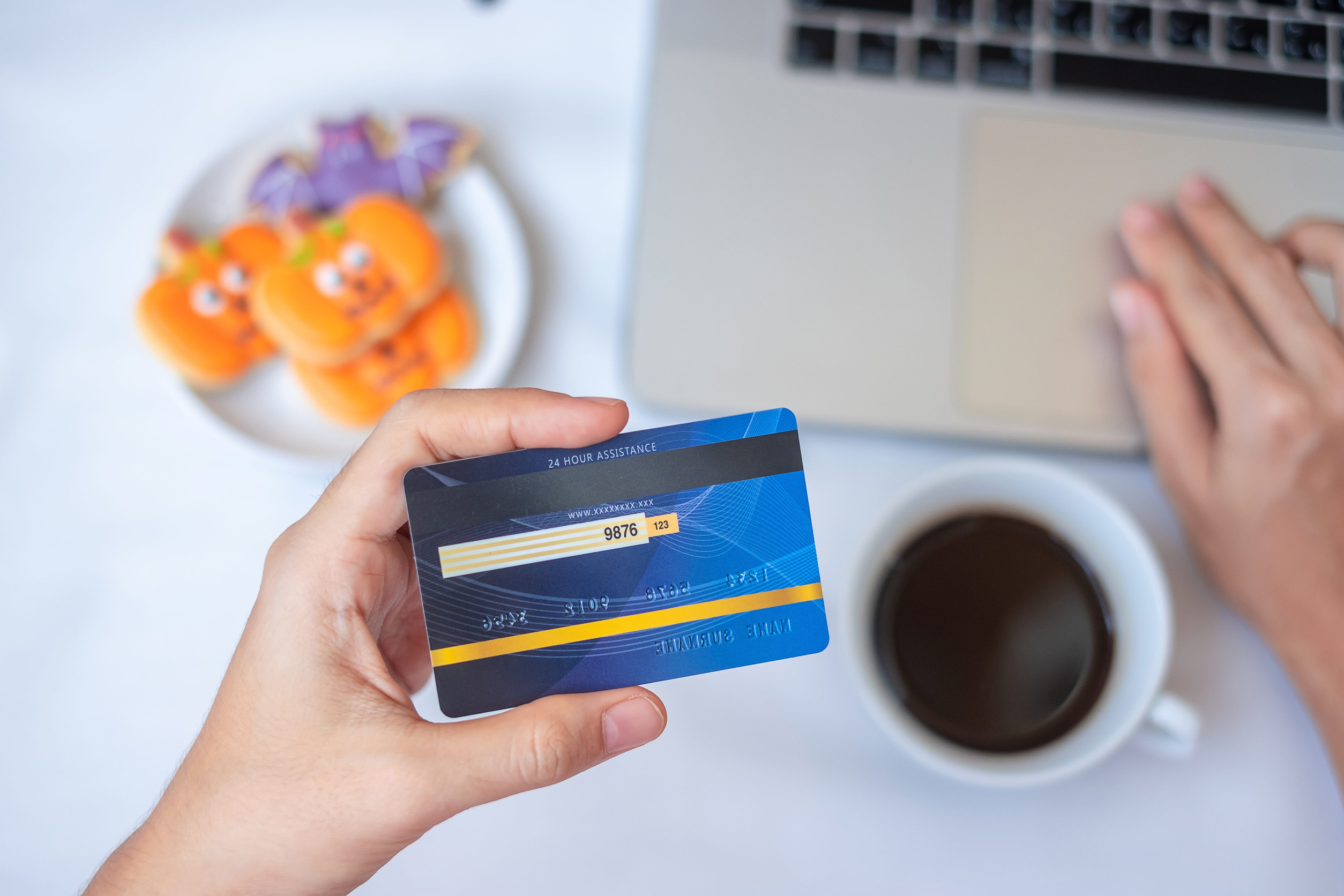 Hand holding credit card for online shopping on laptop during eating Halloween Cookies and coffee. Happy Halloween, Hello October, fall autumn, Festive, party and holiday concept
