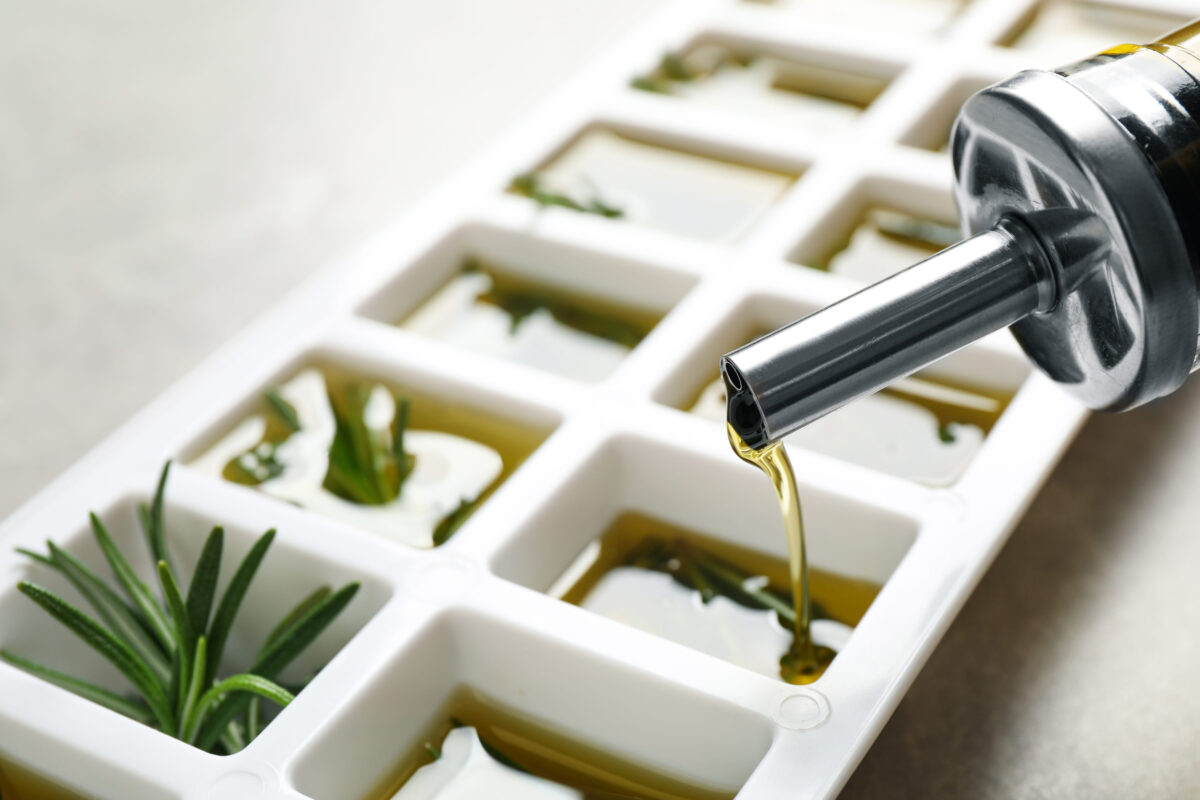 Pouring olive oil into ice cube tray with fresh rosemary at gray table, closeup