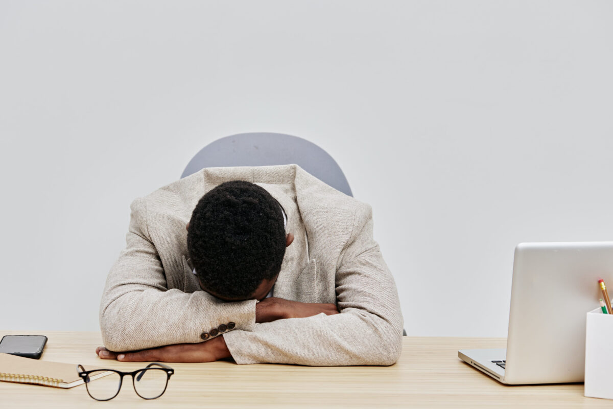 Black and white photo of a tired man resting his head on his arms at a desk with a laptop, glasses, and a smartphone, conveying fatigue or stress in a modern office environment. Business work concept.