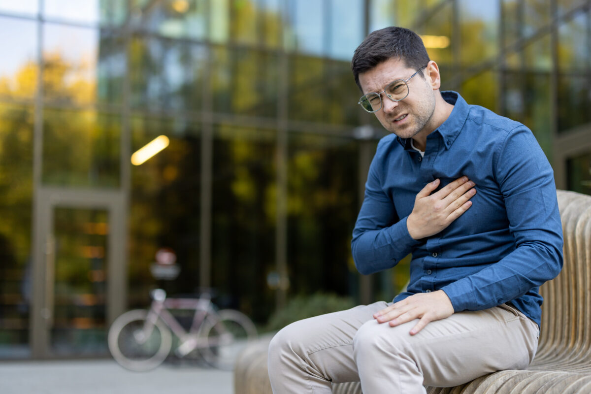 Young man experiencing acute chest pain and discomfort, sitting on a bench outside a modern building, showing symptoms of a potential heart attack or cardiac emergency