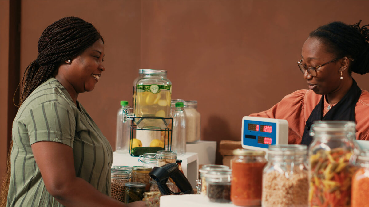 Vendor using electronic scale at checkout counter, selling organic homemade produce for vegan client at local supermarket. African american merchant putting natural fresh merchandise in a paper bag.
