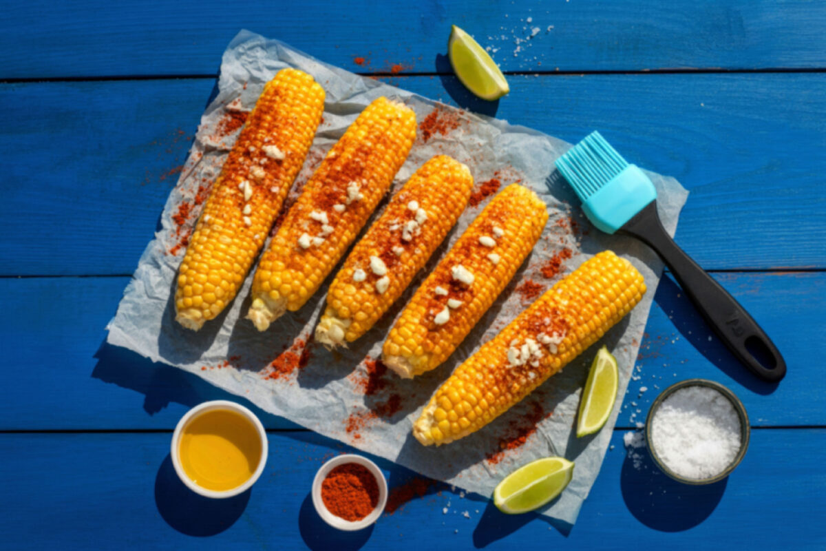 Four grilled corn cobs seasoned with spices and accompanied by a brush, lime slices, and small containers of butter, spices, and salt on a blue wooden table.