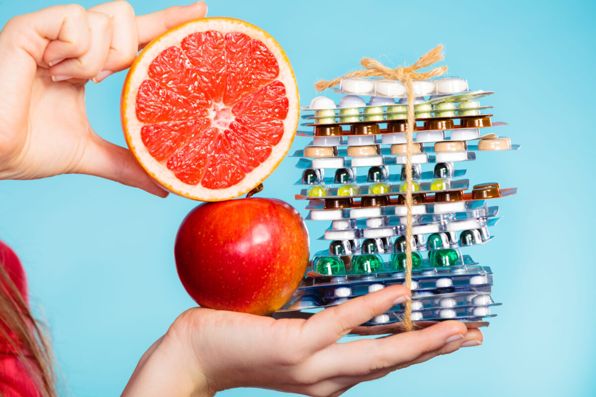 Health and balanced diet concept. Choice between two sources of vitamins - pills or fruits. Closeup female hand holding stack of drugs apple and grapefruit on blue.