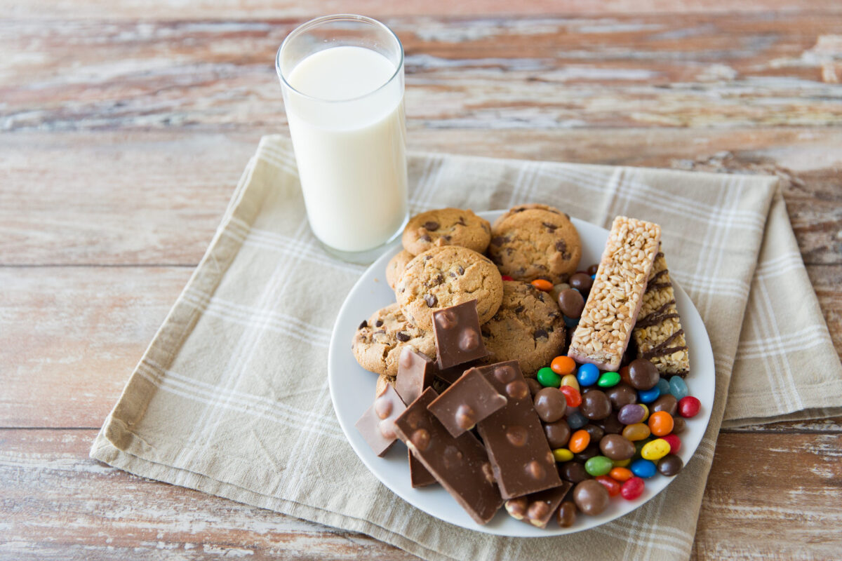 junk food, sweets and unhealthy eating concept - close up of candies, chocolate, muesli and cookies with milk glass on plate
