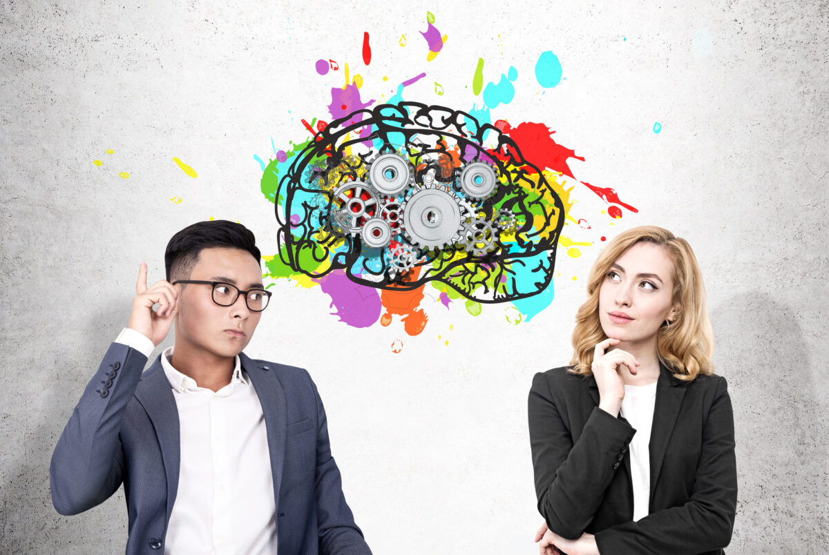Portrait of an Asain businessman and a blond woman standing near a concrete wall with a colorful brain sketch and cogs drawn on it.