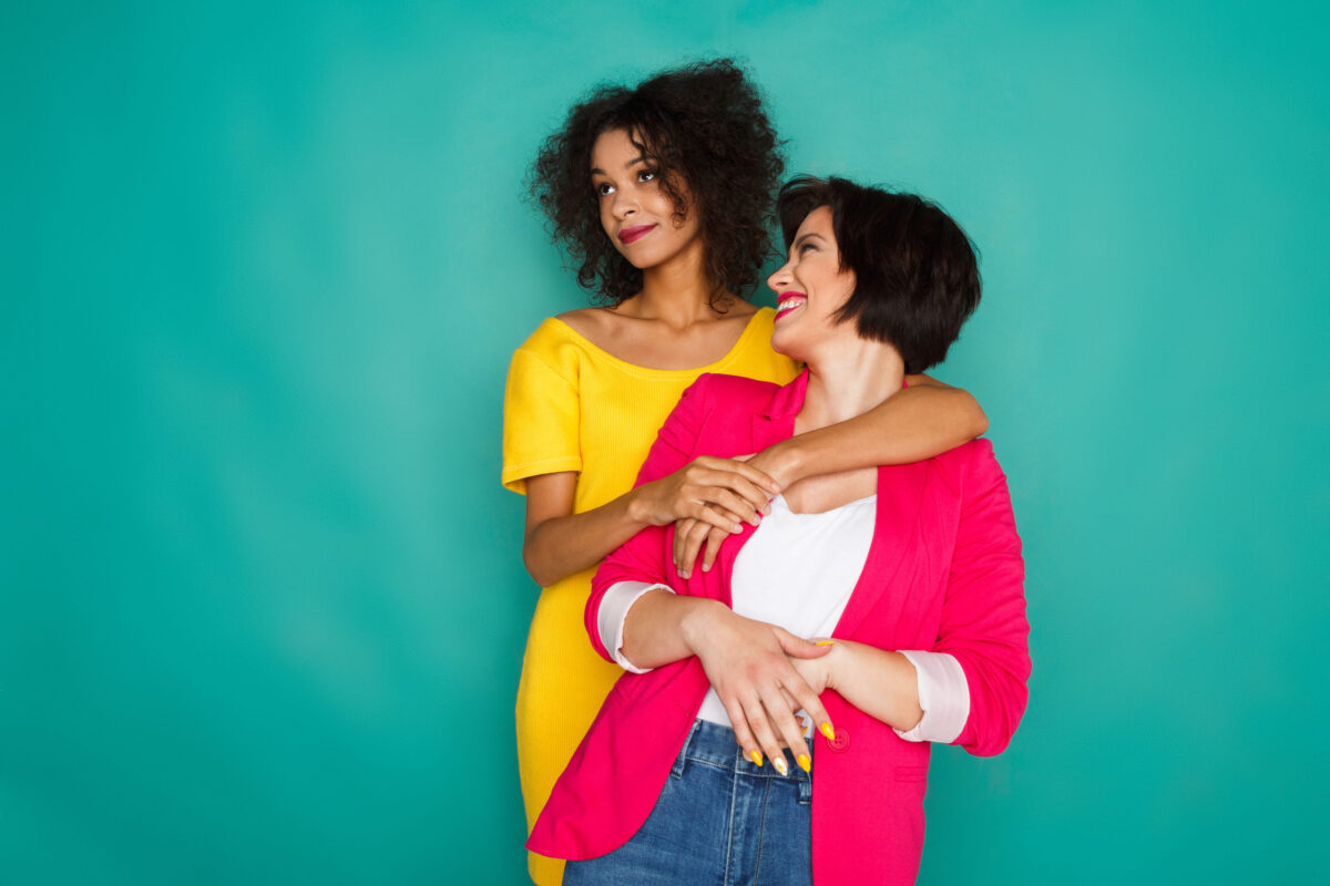 Multiethnic friendship and support concept. Mulatto and caucasian girls in colorful casual clothes hugging and having fun at blue studio background with copy space