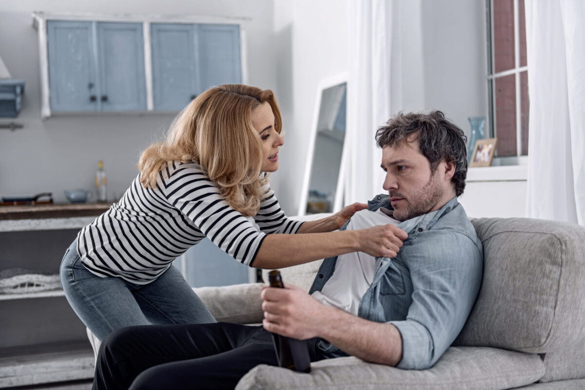 Young expressive caring woman holding her alcoholic husband by the clothes and talking to him while persuading him to give up his awful habit