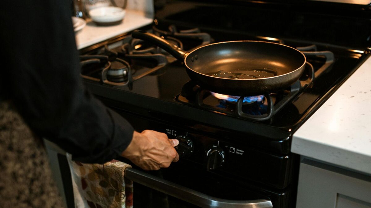 A person in a modern kitchen adjusting the stove dial under a frying pan on a gas burner