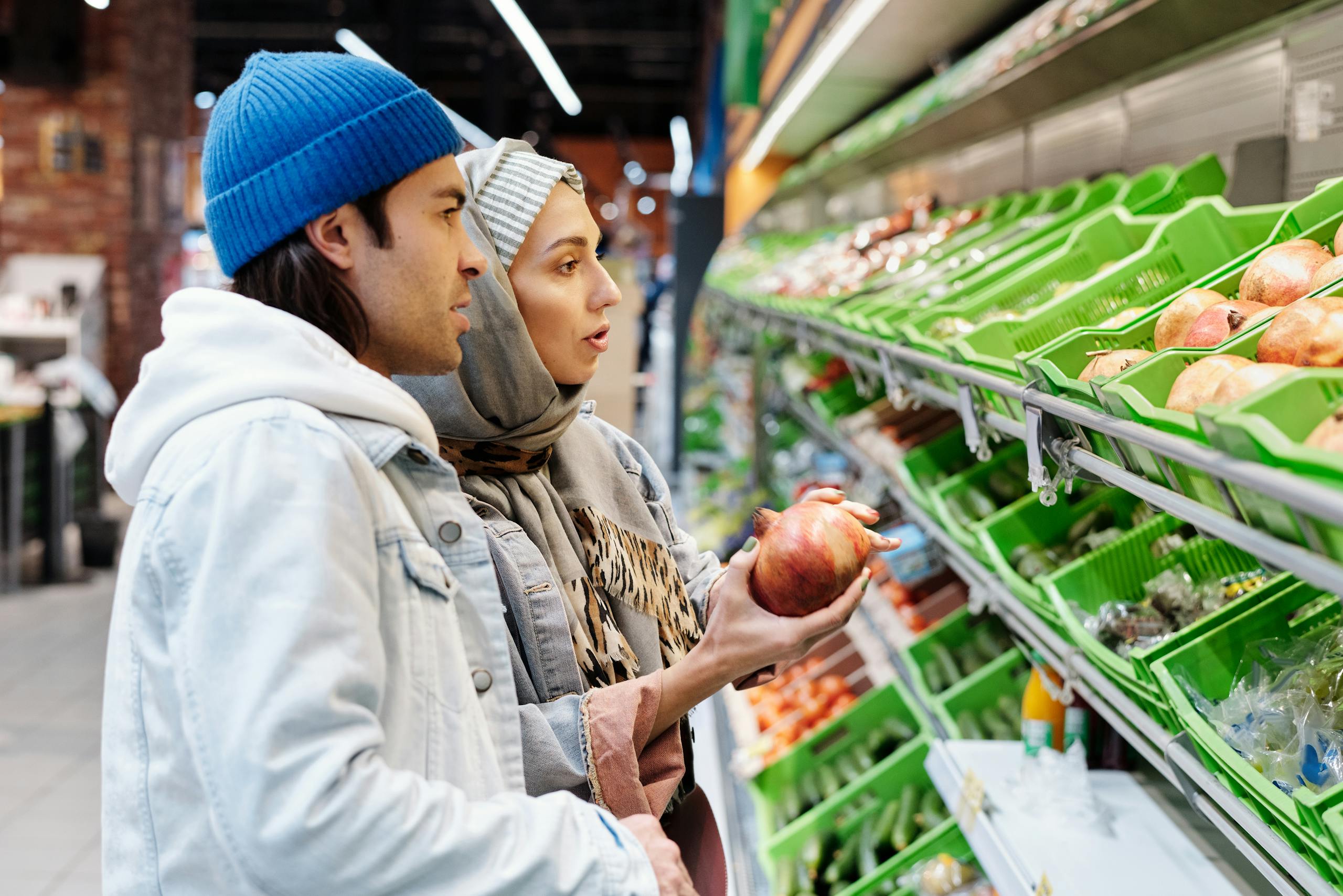 7 Reasons Americans Don’t Believe the Economy Is Fine Anymore 1 A couple selecting pomegranates in a grocery store aisle filled with fresh produce.