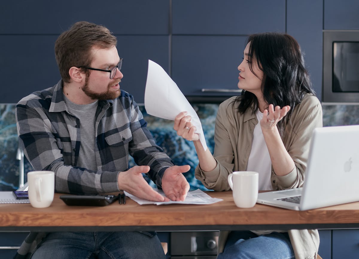 7 Things That Can Destroy a Marriage or Long-Term Relationship, and How to Avoid Them 1 A couple sits at a table, discussing finances with papers and a laptop, looking concerned.