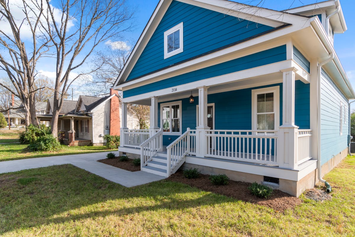 A cozy blue suburban house with a white front porch and lush surroundings on a clear day.