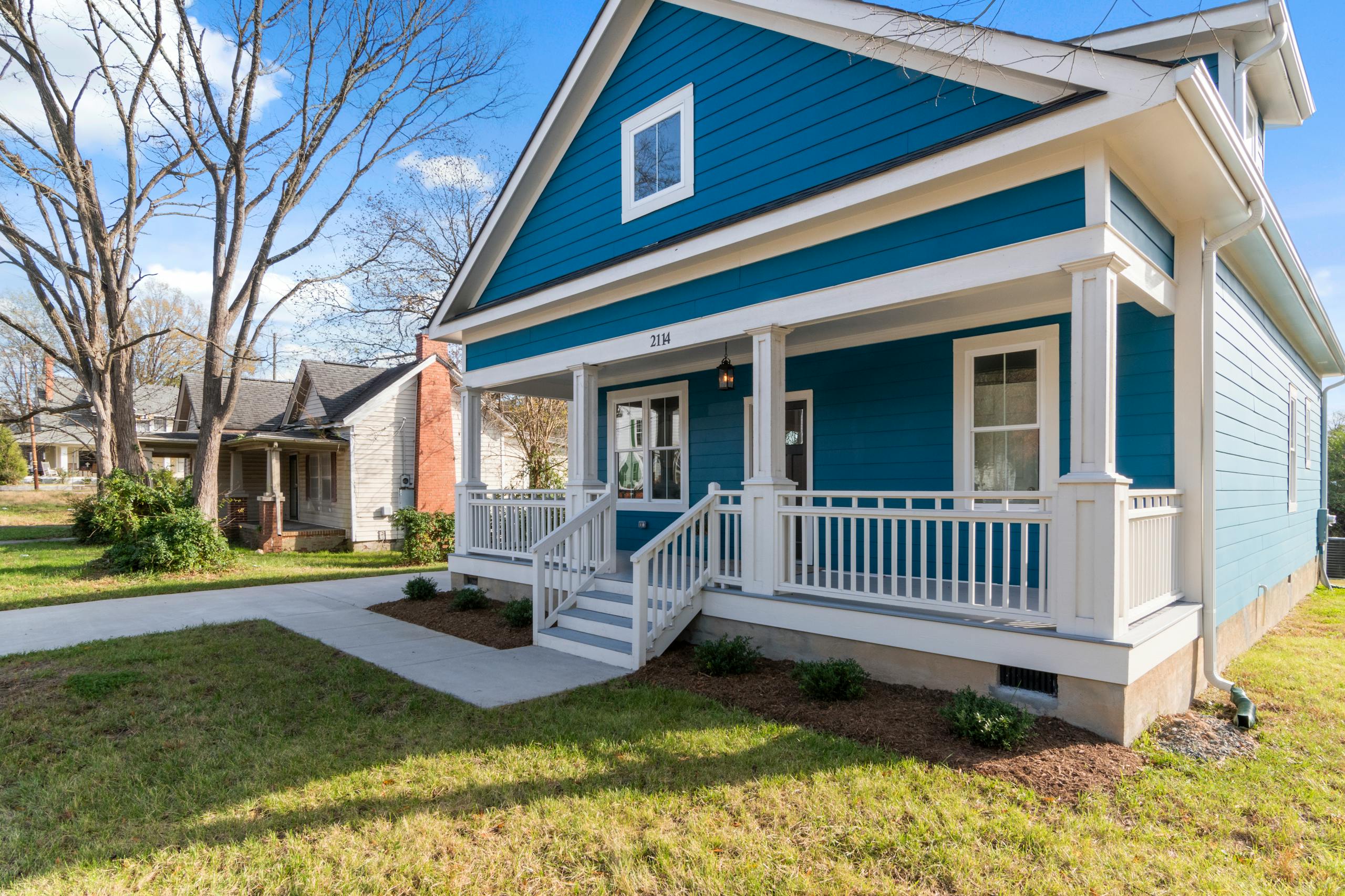 A cozy blue suburban house with a white front porch and lush surroundings on a clear day.