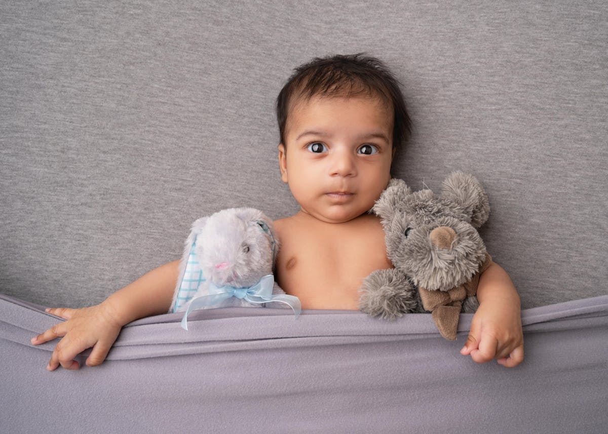 A cute baby lies on a grey blanket with stuffed bunny rabbit and teddy bear, looking playful and cozy.