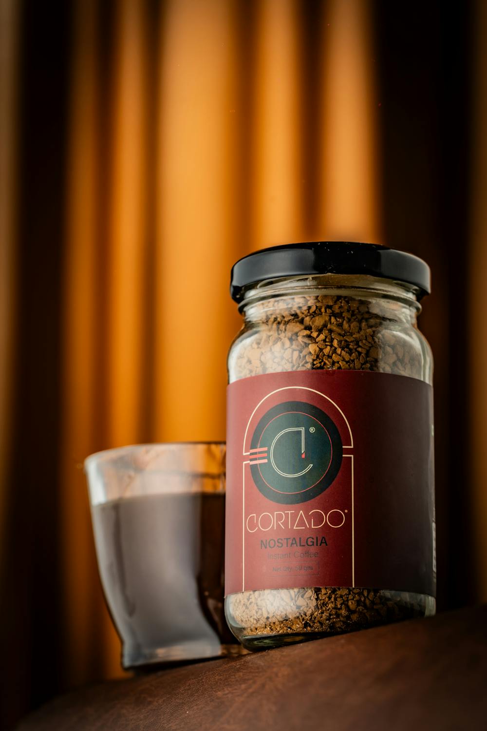 A jar of instant coffee labeled 'Cortado Nostalgia' beside a cup on a wooden surface with warm lighting.