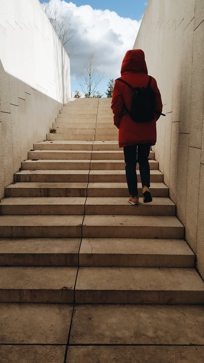 A lone figure in a red coat ascends an outdoor stone staircase under a clear sky.