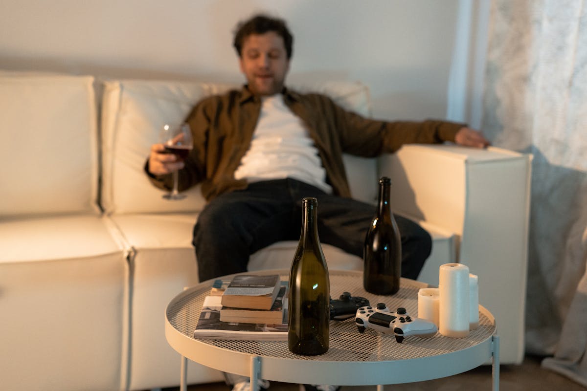 8 Disturbing Truths About Getting Drunk 1 A man sits on a couch with wine bottles, a game controller, and books, expressing relaxation or contemplation.
