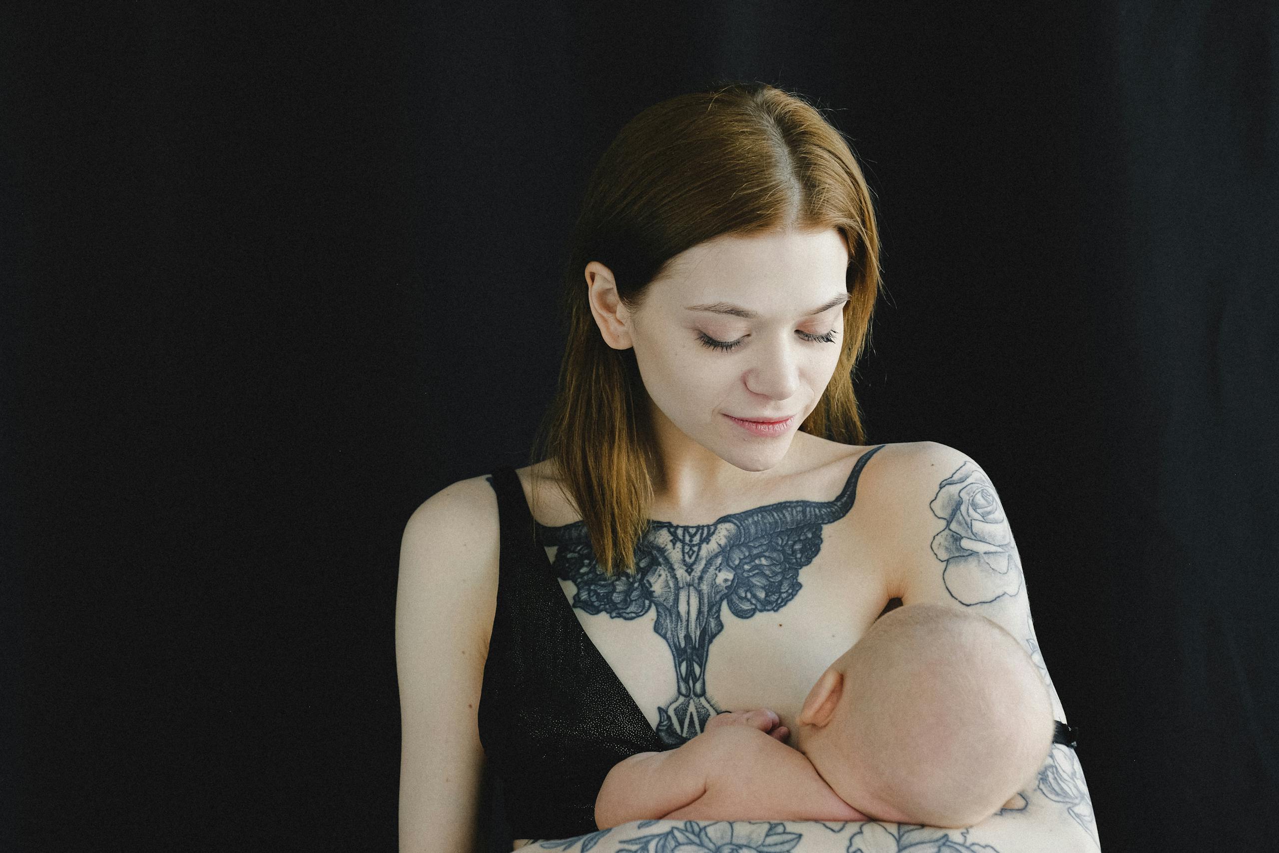 7 Reasons Botox After Pregnancy Can Become a Beauty Decision You Regret 1 A mother with tattoos lovingly breastfeeding her infant against a dark backdrop.