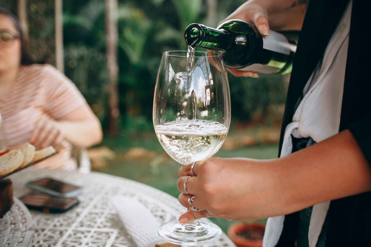 A person pours white wine into a glass during a gathering in a garden setting.