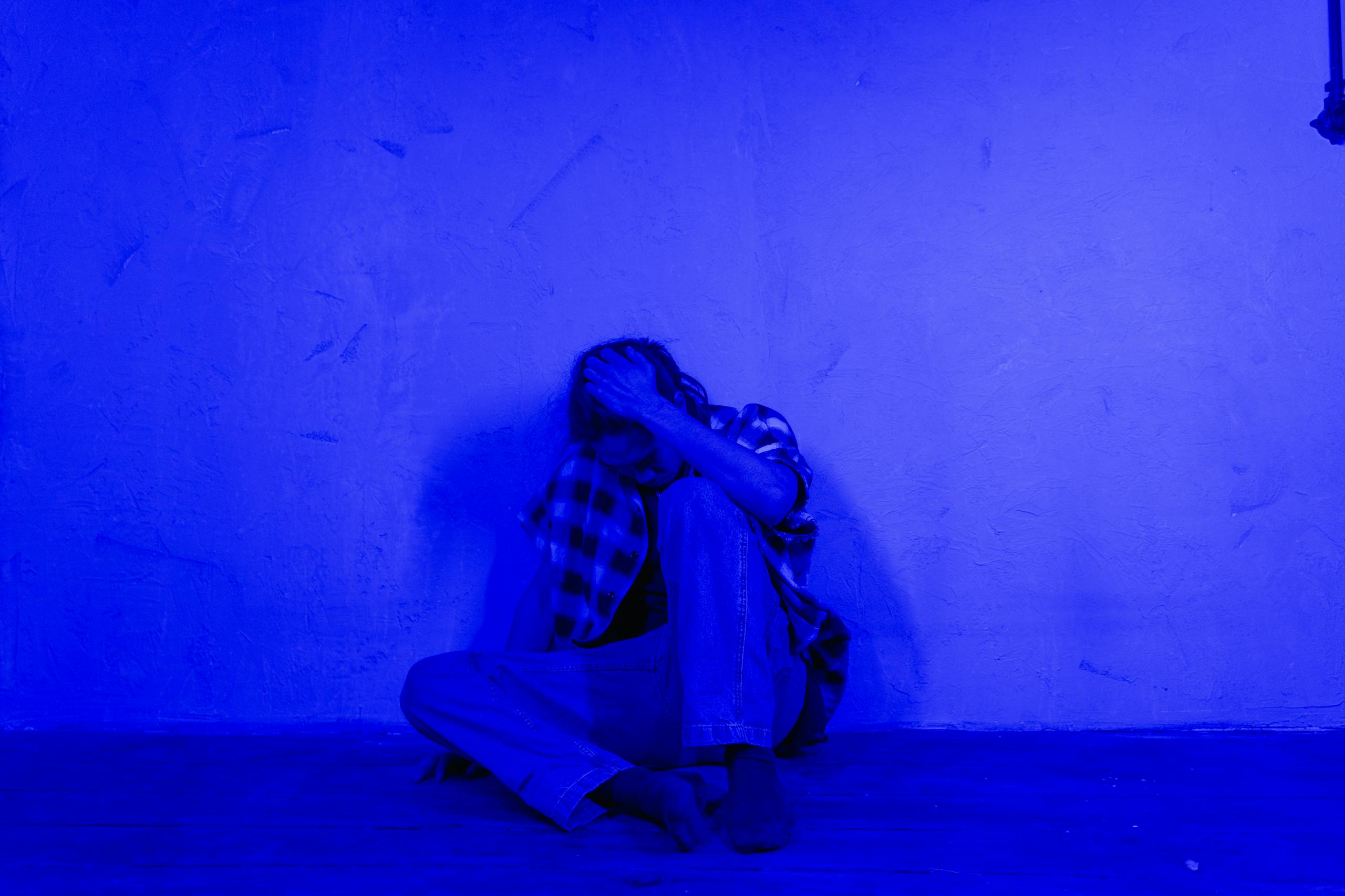 10 Disturbing Reasons Modern Friendships Are Falling Apart in America 4 A person sitting against a wall in a blue-lit room, expressing solitude and contemplation.