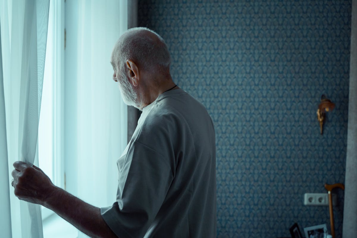 A senior man stands alone, looking out a window with an expression of deep contemplation.