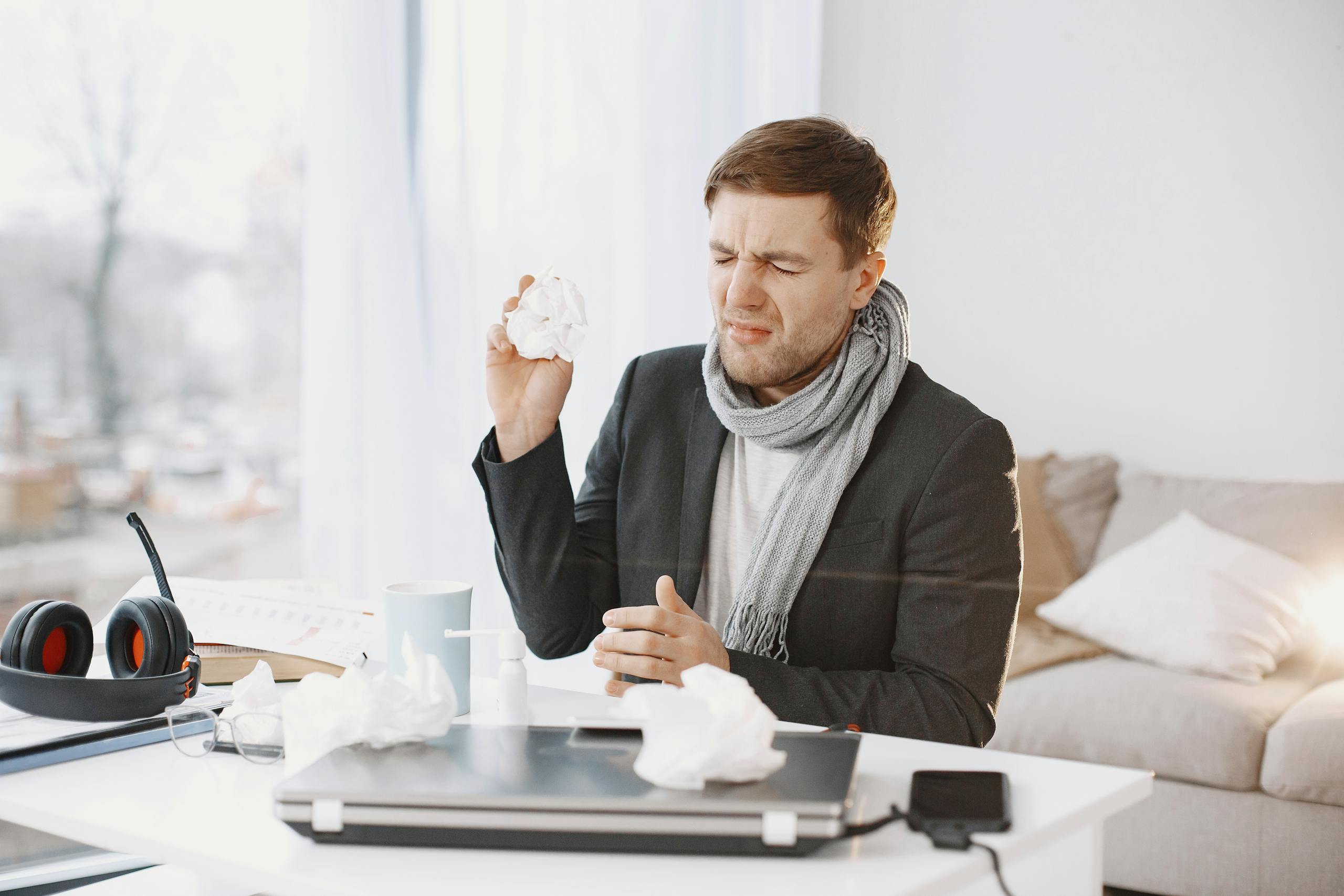 A sick man wearing a scarf sitting at his desk, holding a tissue, displaying signs of discomfort.