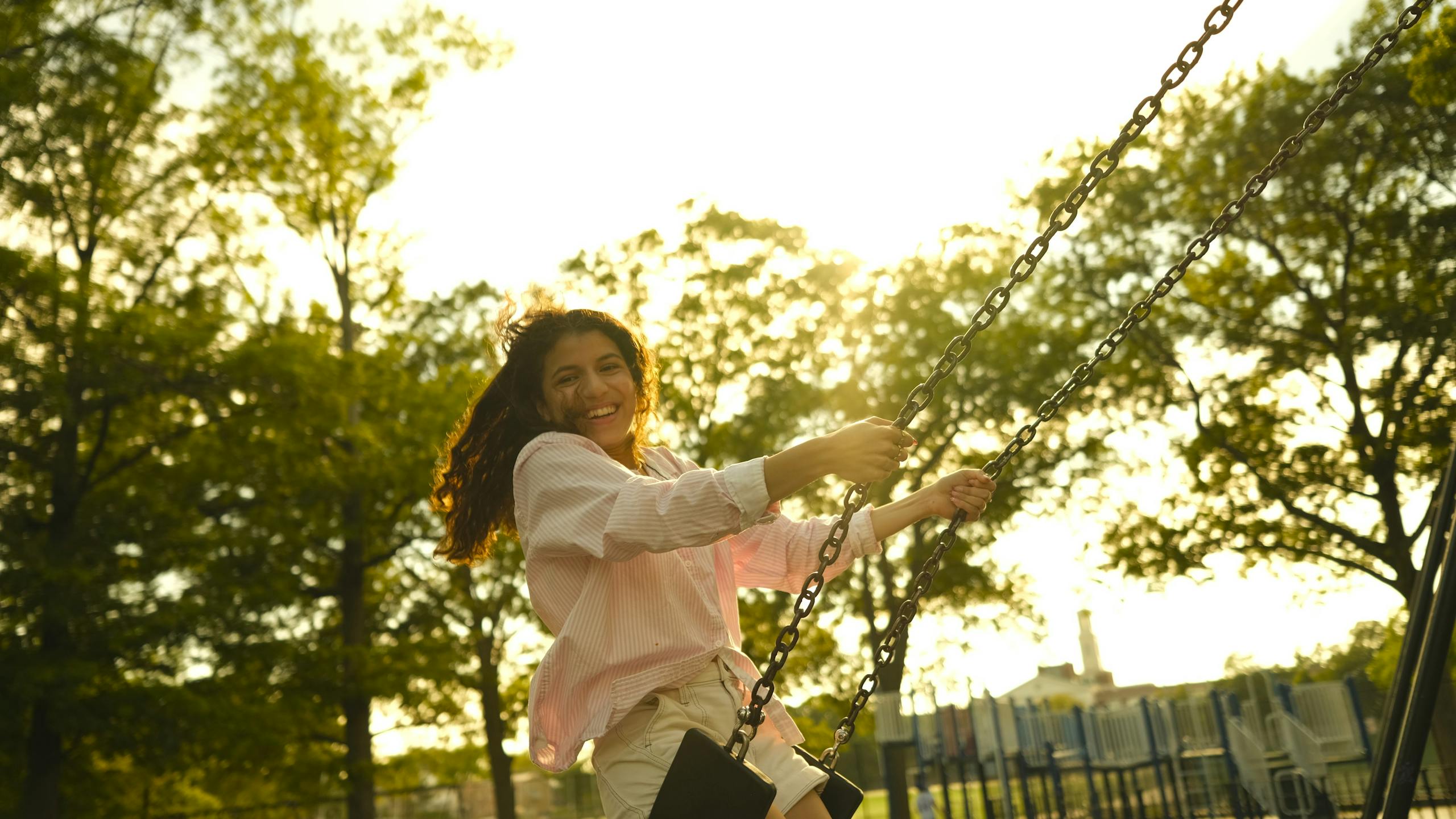 A smiling woman enjoys swinging at a sunny playground, capturing a moment of joy and leisure.