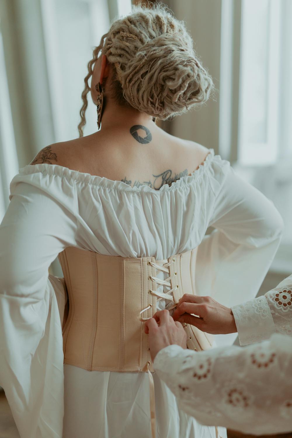A stylist tightens a vintage corset on a tattooed woman's back, highlighting elegance.
