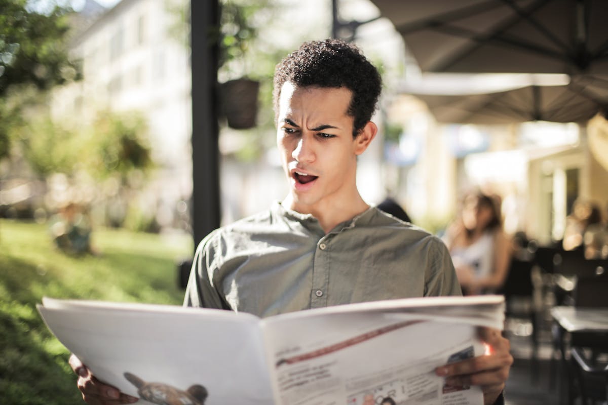 A surprised man in casual wear reads a newspaper while sitting outdoors, expressing shock.