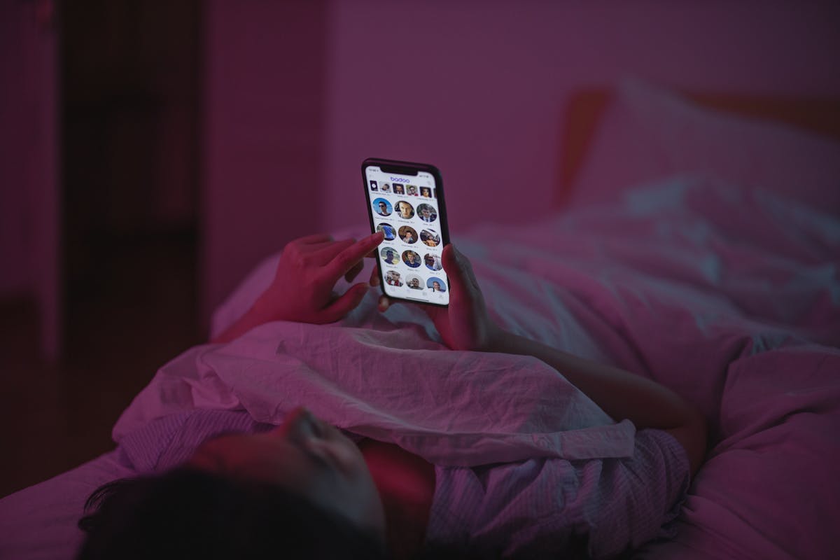 A woman relaxes in bed at night, browsing on her smartphone under pink ambient lighting.