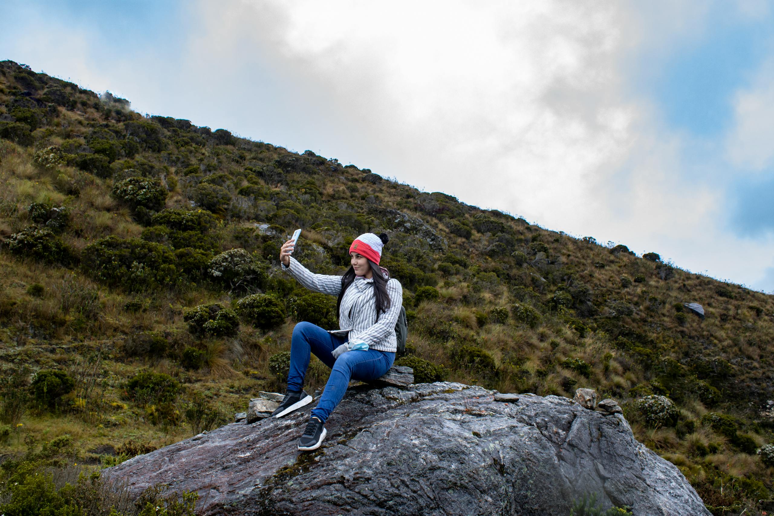 A woman sits on a rocky hilltop, capturing a selfie amidst the lush Colombian landscape.