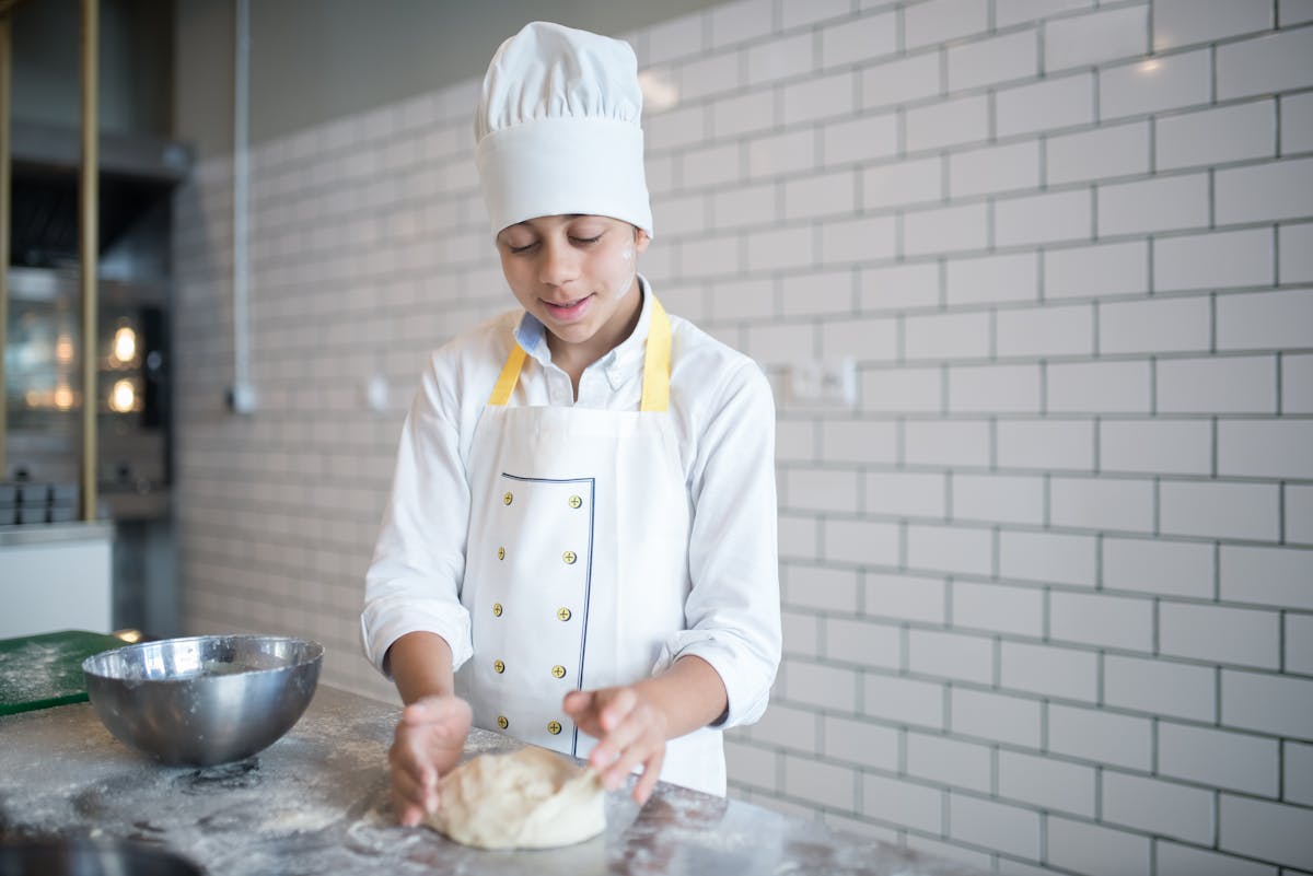 A young chef in a toque kneads dough on a flour-dusted kitchen counter.