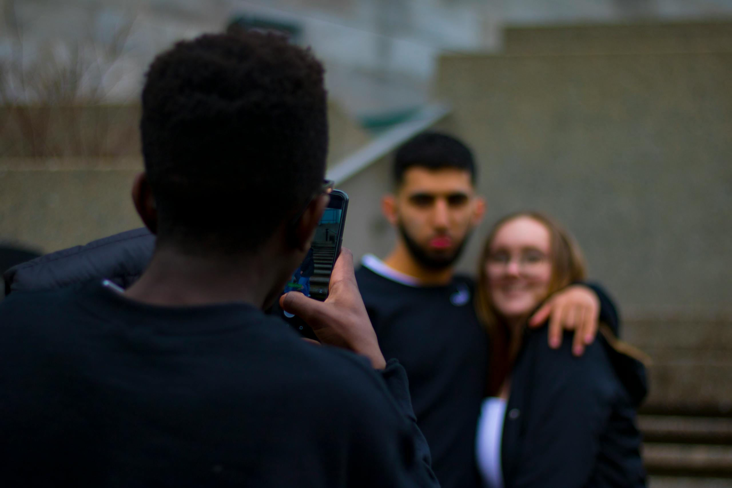 A young couple smiles as their friend takes a photo with a smartphone in an urban outdoor setting.