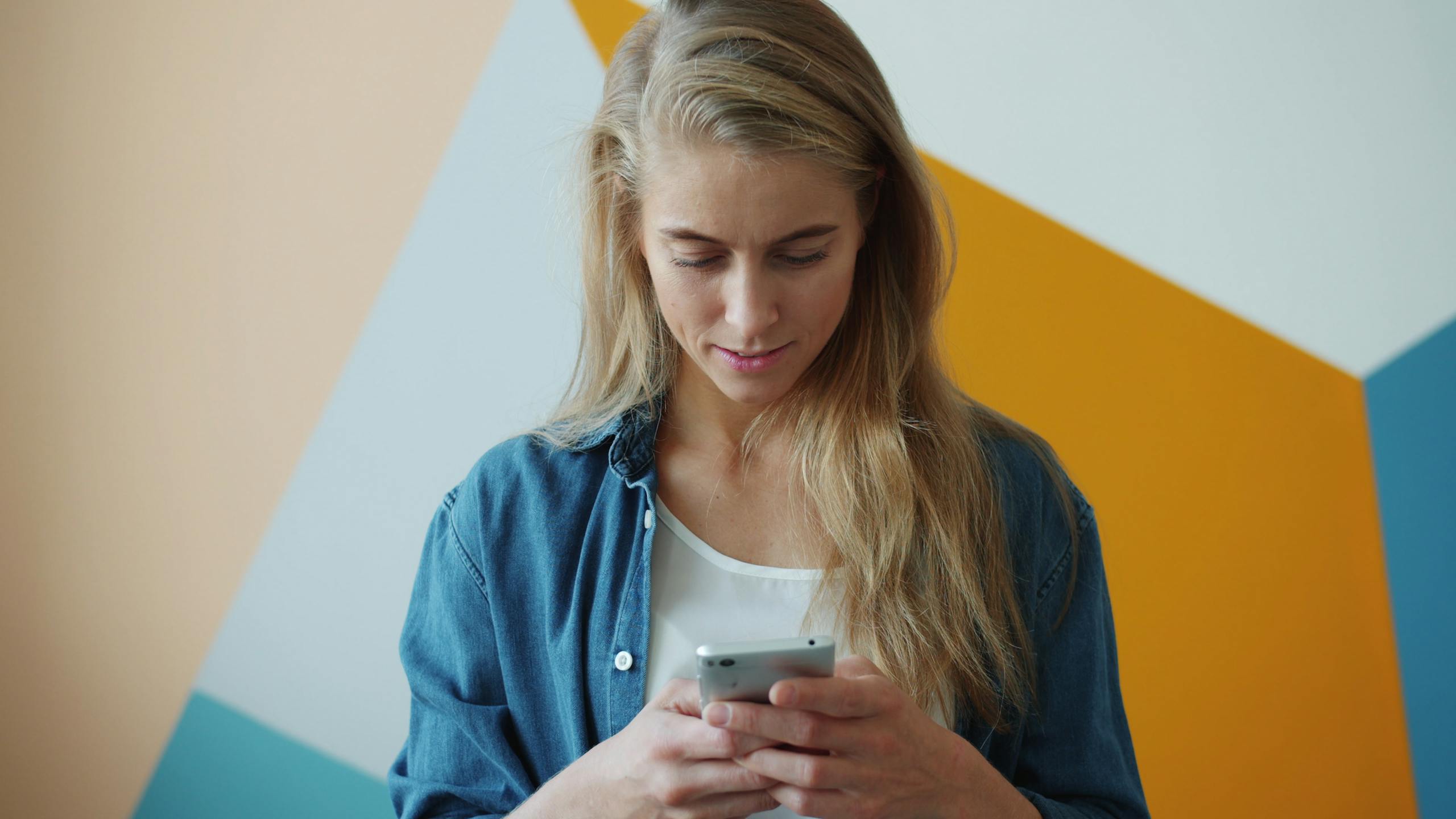 A young woman in a denim shirt uses a smartphone against a geometric colorful wall.