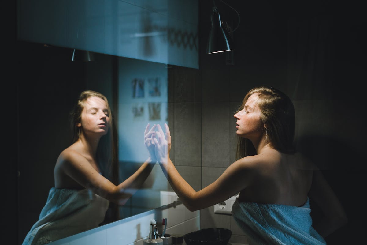 8 Surprising Things You Don’t Realize Are Making You Smell Bad 3 A young woman wrapped in a towel touches the mirror in a contemporary bathroom setting.