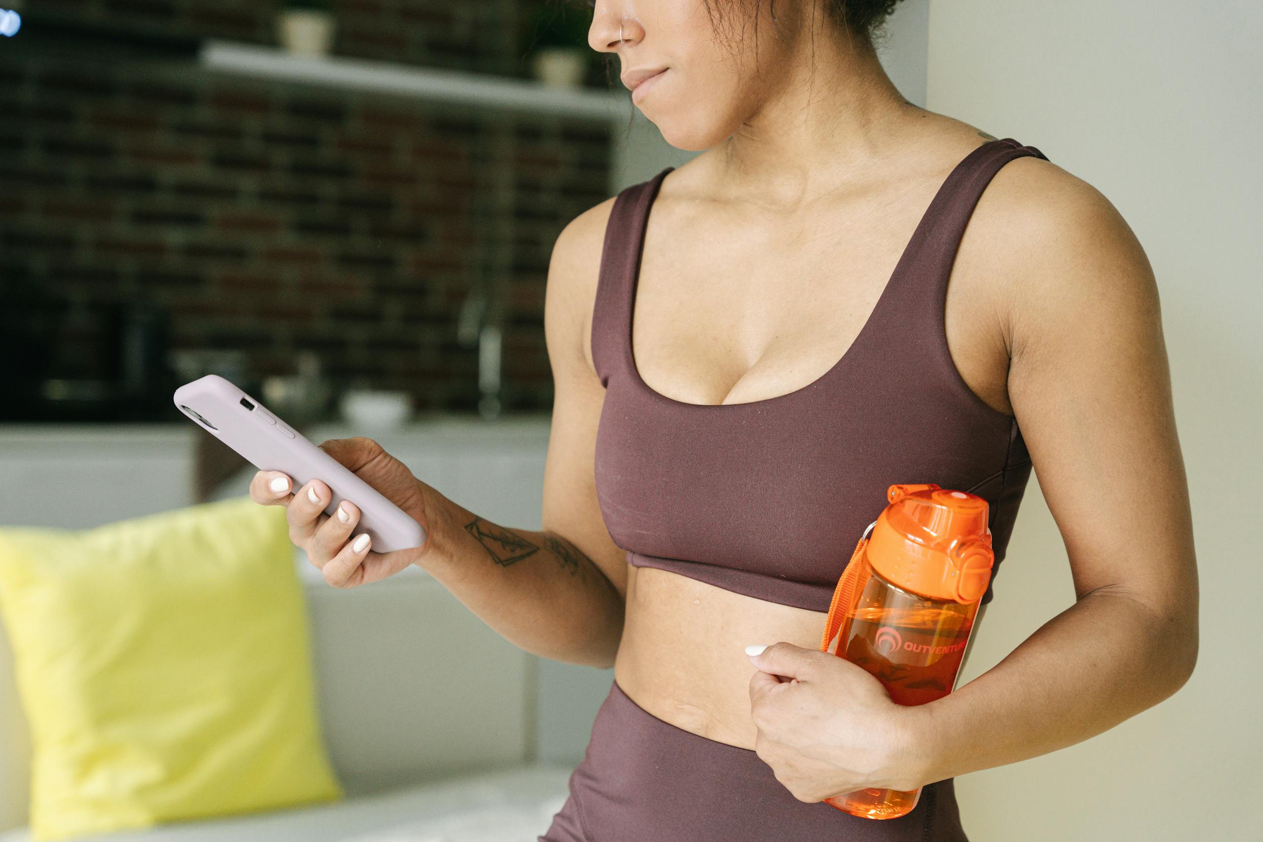 Active woman in a sports bra using a smartphone indoors, holding a water bottle.