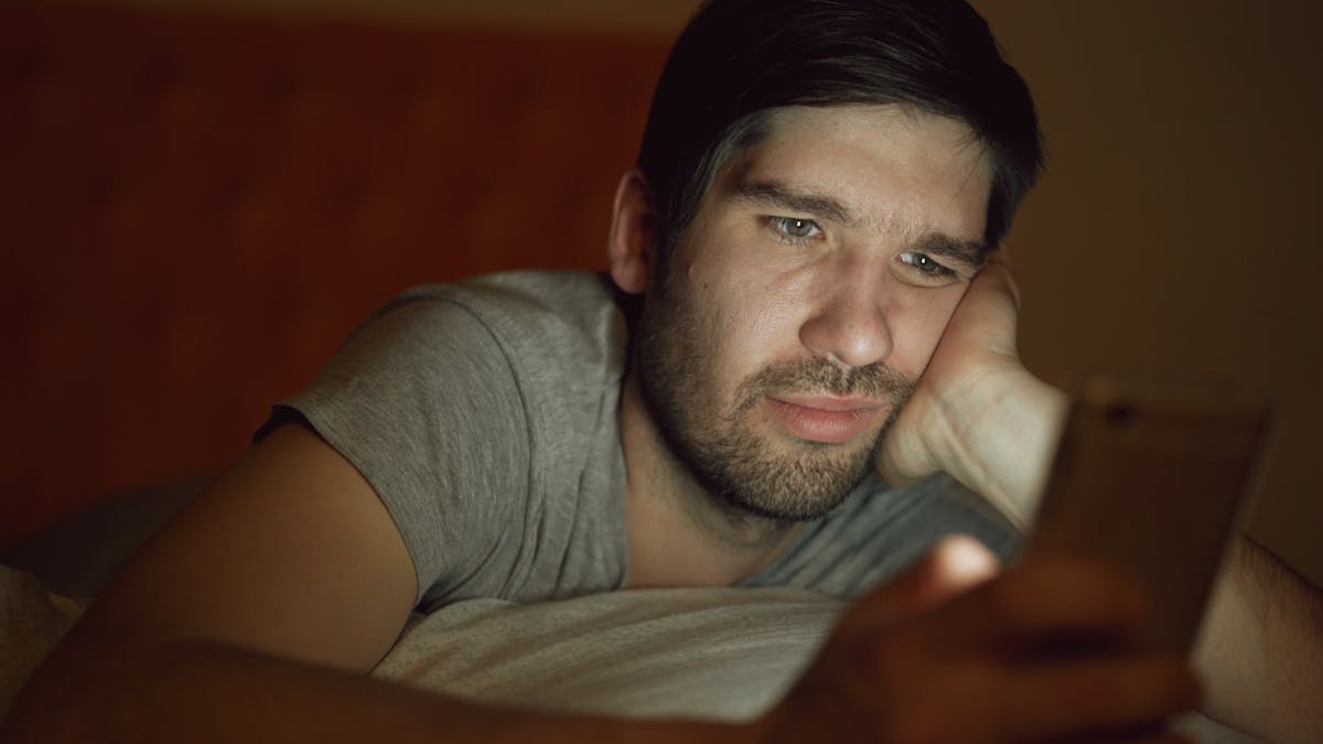 An adult man in a dim room looking at a smartphone with a thoughtful expression.