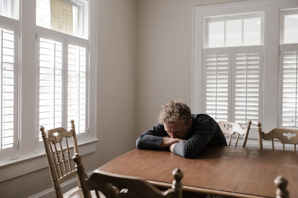 8 Emotional Dangers of Relying Too Much on AI 3 An adult man with emotions of sadness and anxiety sitting alone at a wooden table near windows.
