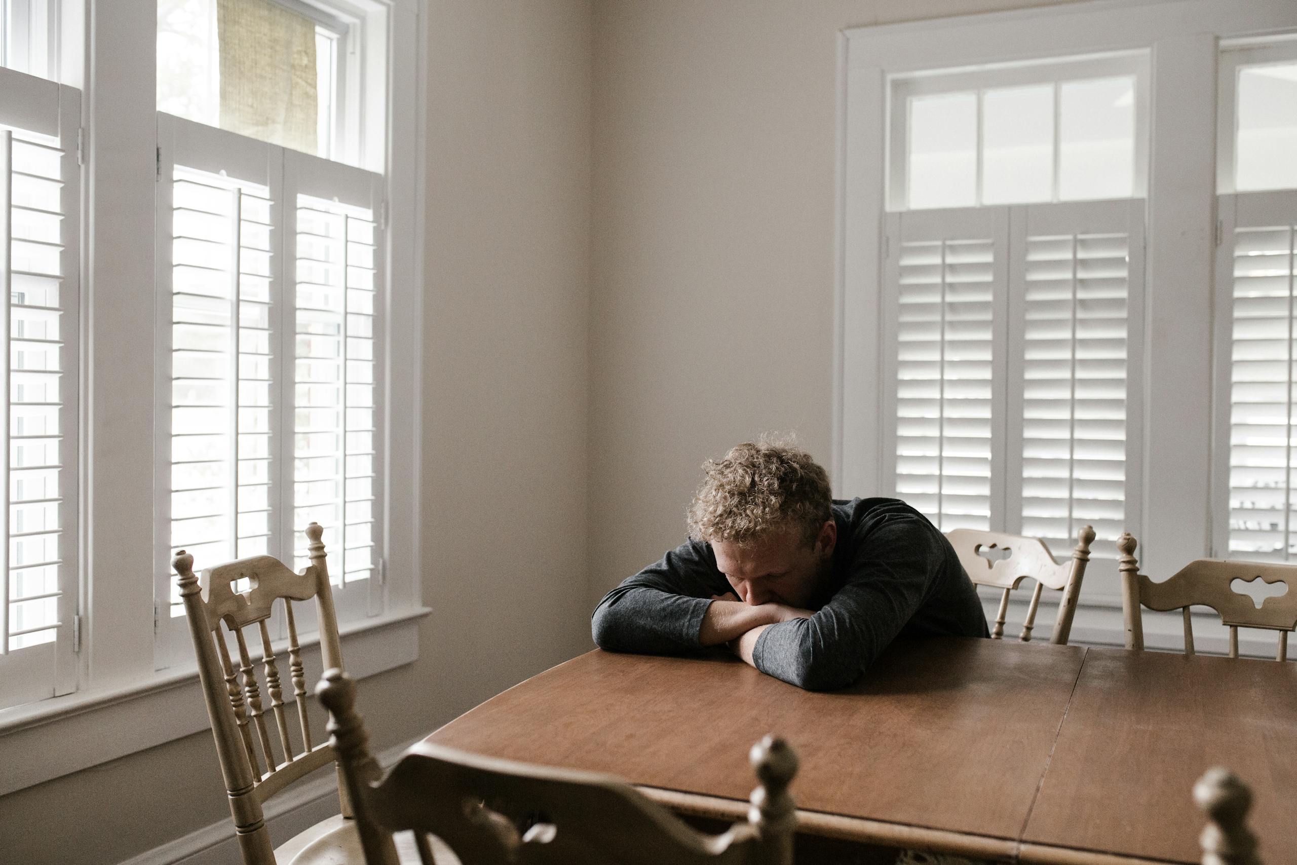 10 Disturbing Reasons Americans Are More Lonely Than They Look Online 4 An adult man with emotions of sadness and anxiety sitting alone at a wooden table near windows.