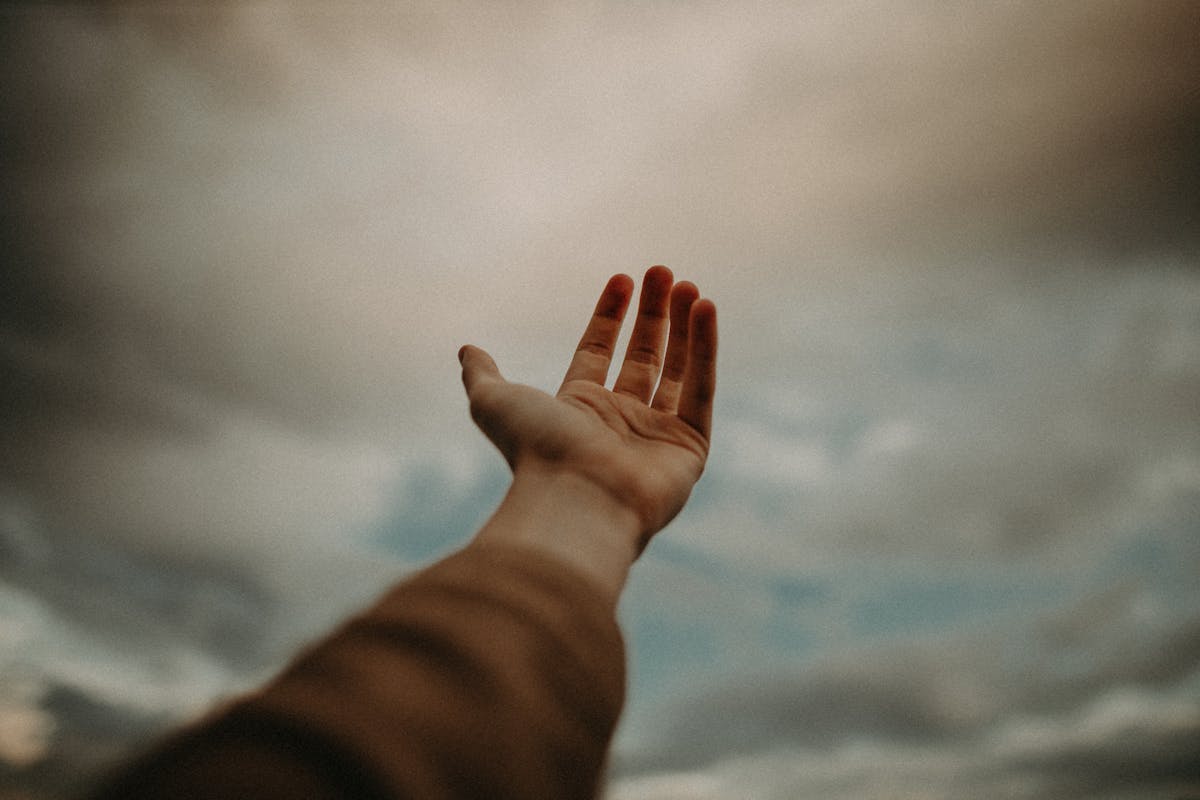 An artistic shot of a hand reaching upward toward a dramatic cloudy sky.