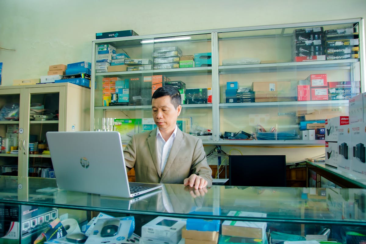 6 Harsh Truths About America’s New Trade War That Consumers Feel First 4 An Asian man using a laptop in an electronics shop with various devices on display.
