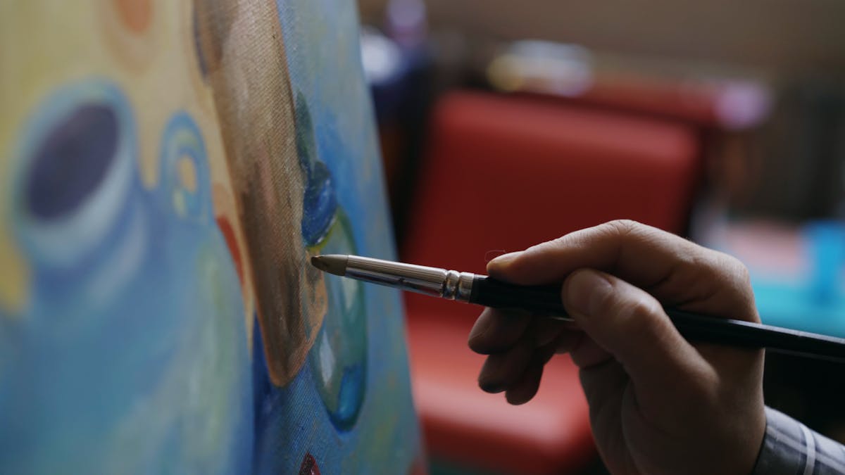 Artist's hand skillfully paints a colorful canvas indoors with a focus on the brush.