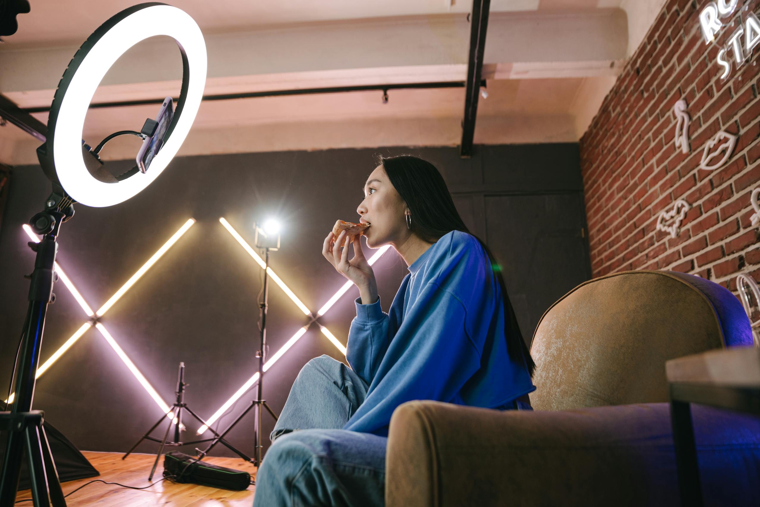 Kai Trump’s “My House” White House Comment Backfired Because American Taxpayers Know Who Really Pays For It 2 Asian woman content creator eating pizza while recording a video indoors with ring light.